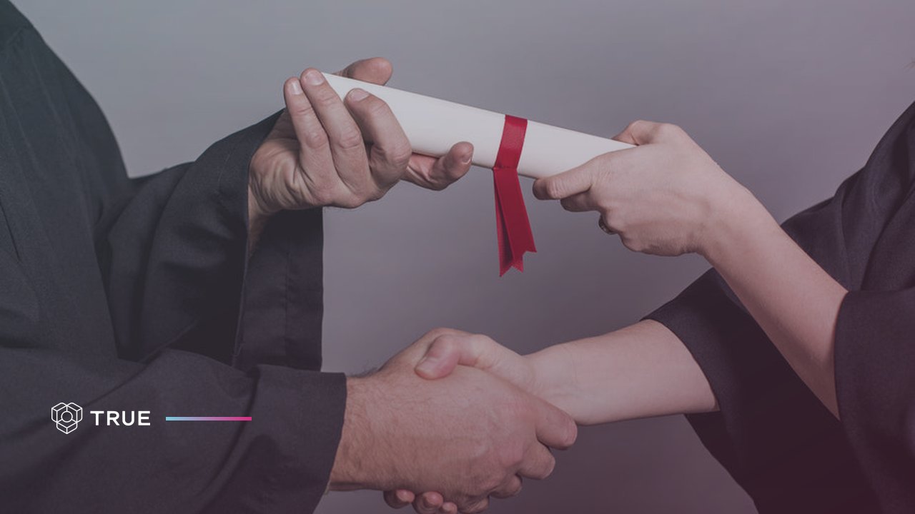 Two people exchanging a rolled diploma with a red ribbon over a handshake